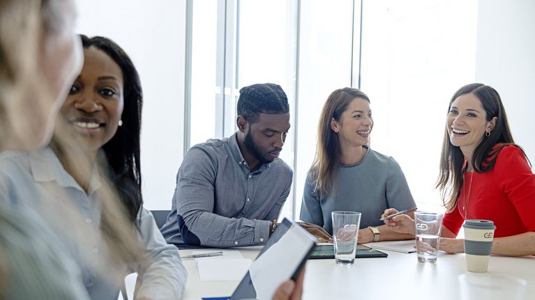 Some employees sit at a large table in a meeting room and talk to each other