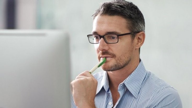 A man sits in front of a computer screen and holds a pen in his hand
