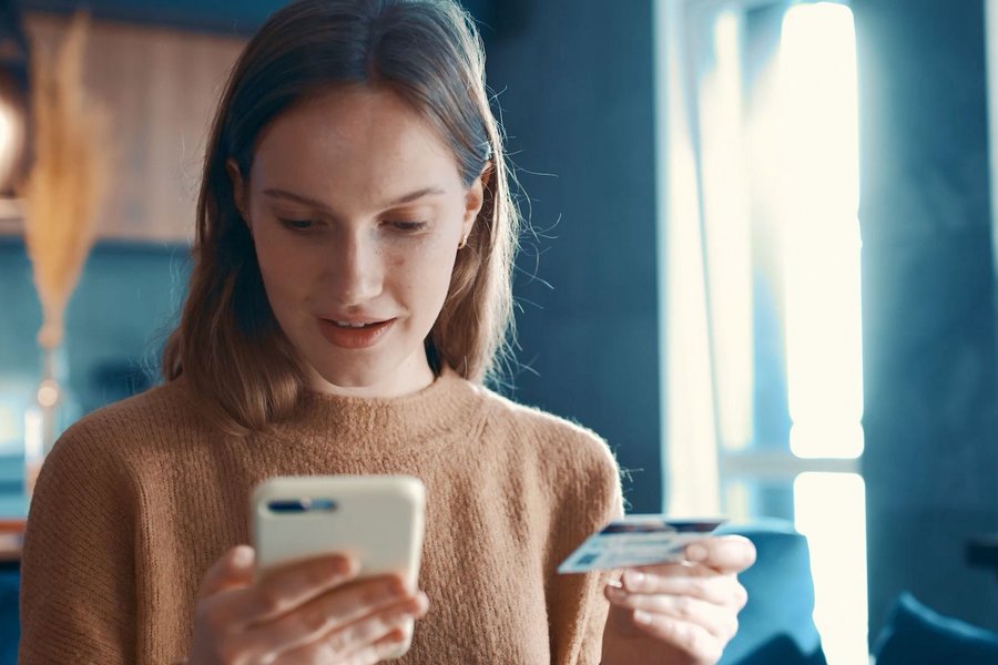 A woman sits in the kitchen holding a smartphone in one hand and a credit card in the other