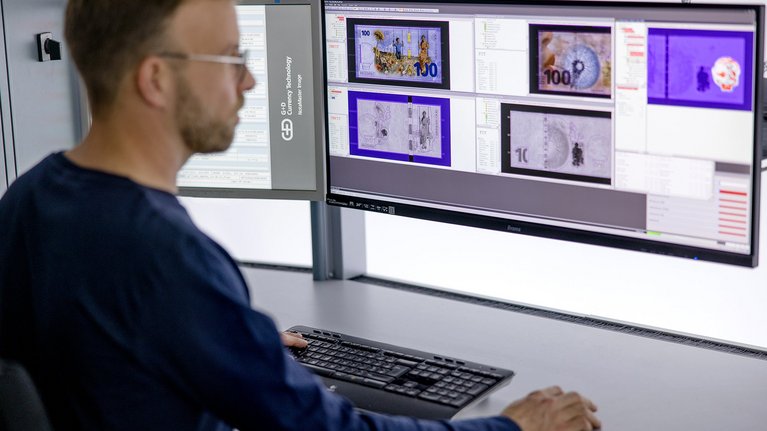 A person is working at a desk with two large computer monitors displaying images of currency notes