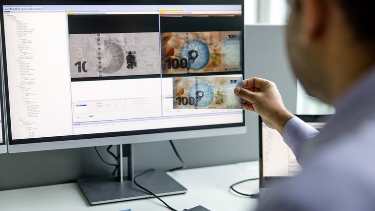 A person is examining a 100 currency note in front of a computer screen displaying magnified images of the same note.