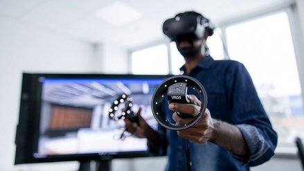 A professional using a virtual reality headset and controllers, standing in an office environment with a monitor displaying a virtual simulation in the background.
