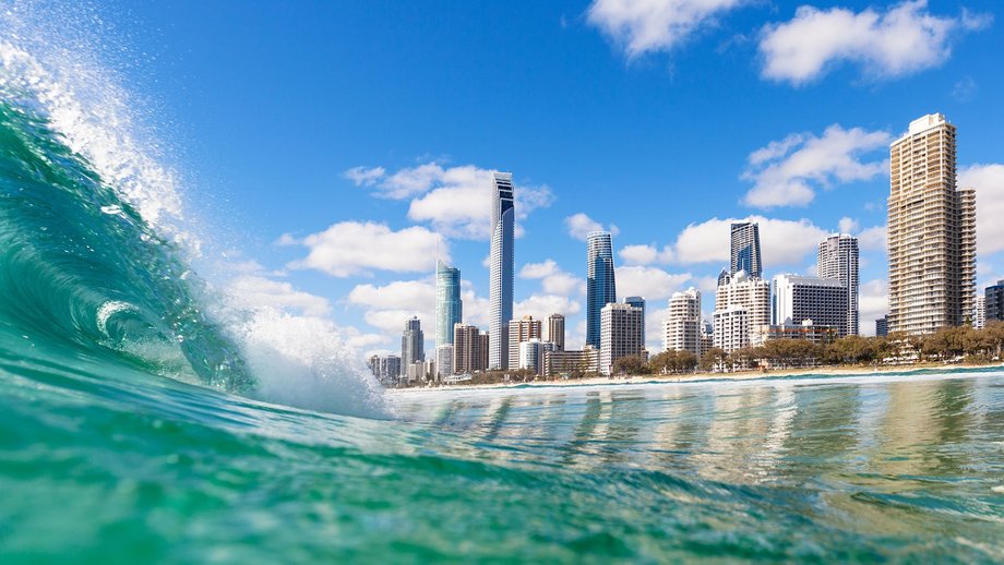 A large ocean wave with a city skyline in the background under a bright blue sky.