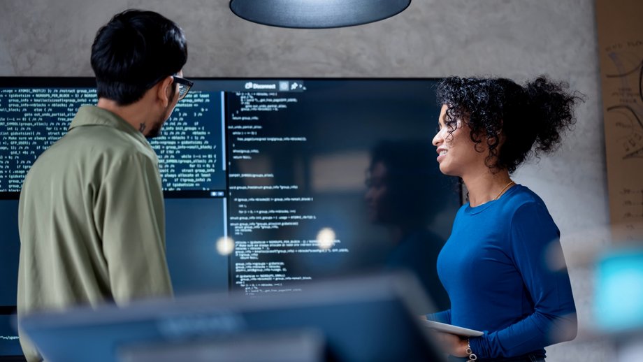 Two developers discuss AI generated code on a large screen in a modern office.