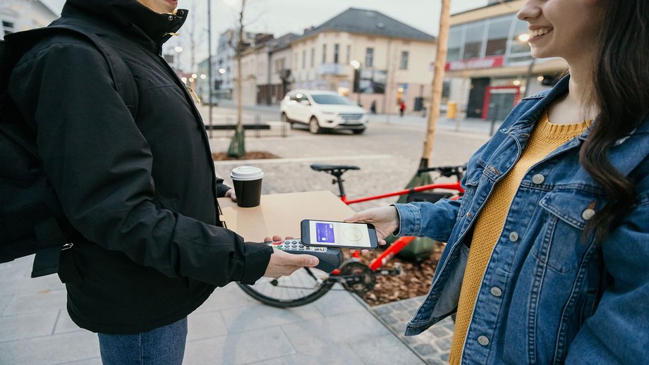 A woman makes a contactless payment with her smartphone to pay a delivery worker for a pizza and coffee