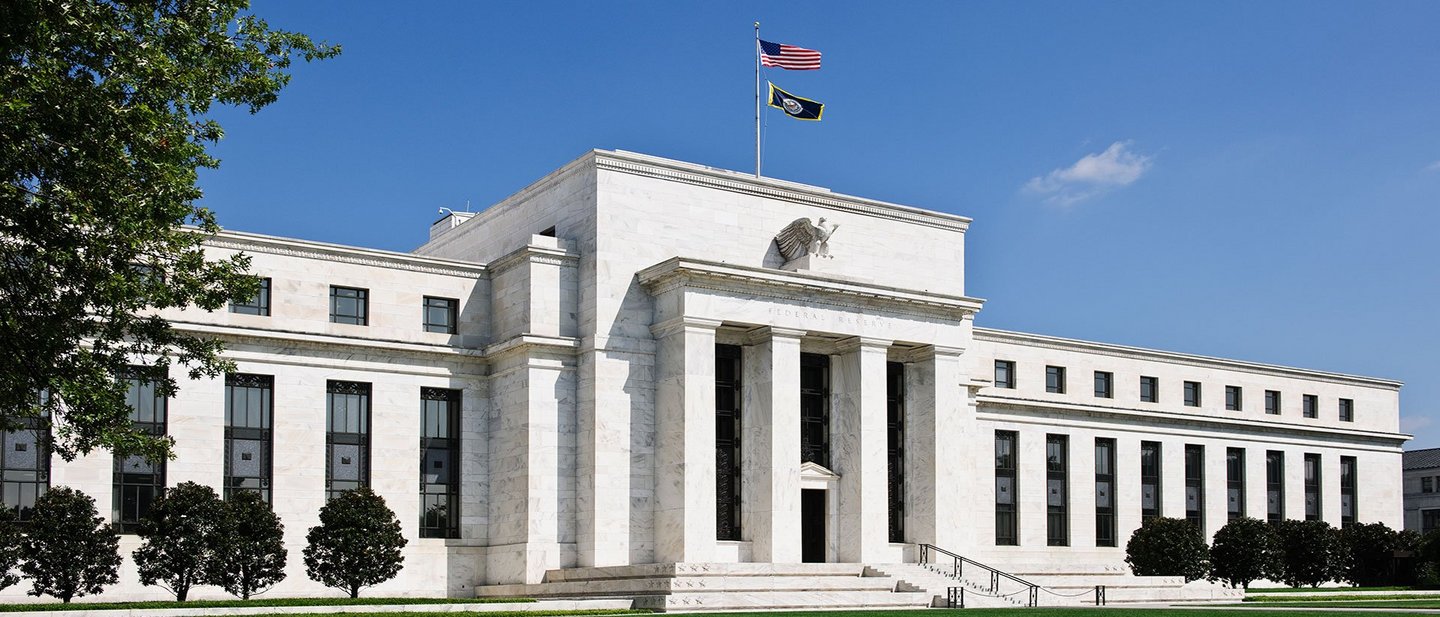 The main entrance of the US Federal Reserve, surrounded by trees, with an American flag hoisted