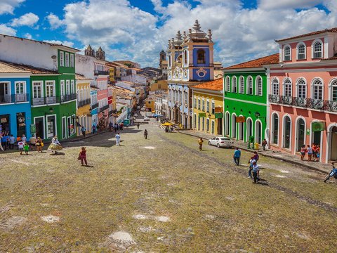 A vibrant street scene in Pelourinho, Salvador, Brazil, with colorful colonial buildings and people walking under a bright blue sky.