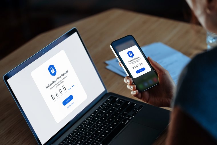 Person holding a smartphone showing a login verification code next to a laptop displaying an authentication screen.