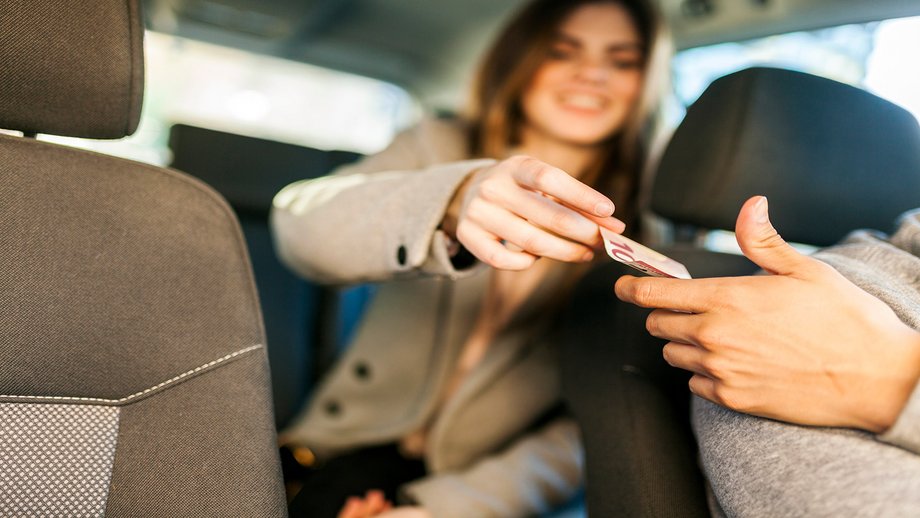 A girl is sitting in the back seat of a car and handing cash to the taxi driver in the front.