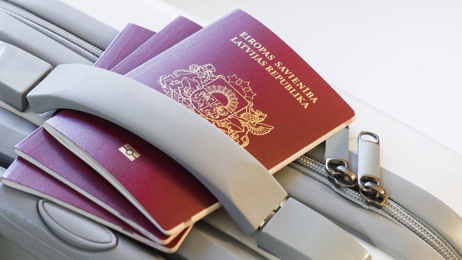 Two Latvian passports rest on a grey suitcase handle, symbolizing travel and mobility.