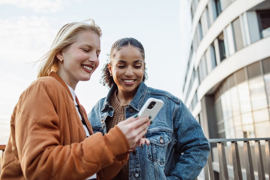 Two young women looking at a white smartphone with futuristic building in the background.