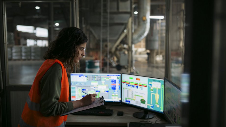 Female technician in a control room looking at computer monitors and making notes on clipboard.