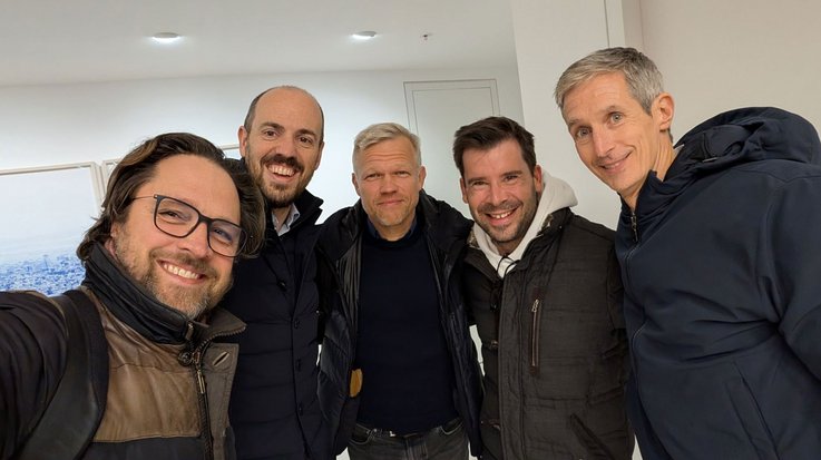 Group of five men standing indoors, dressed in winter clothing.