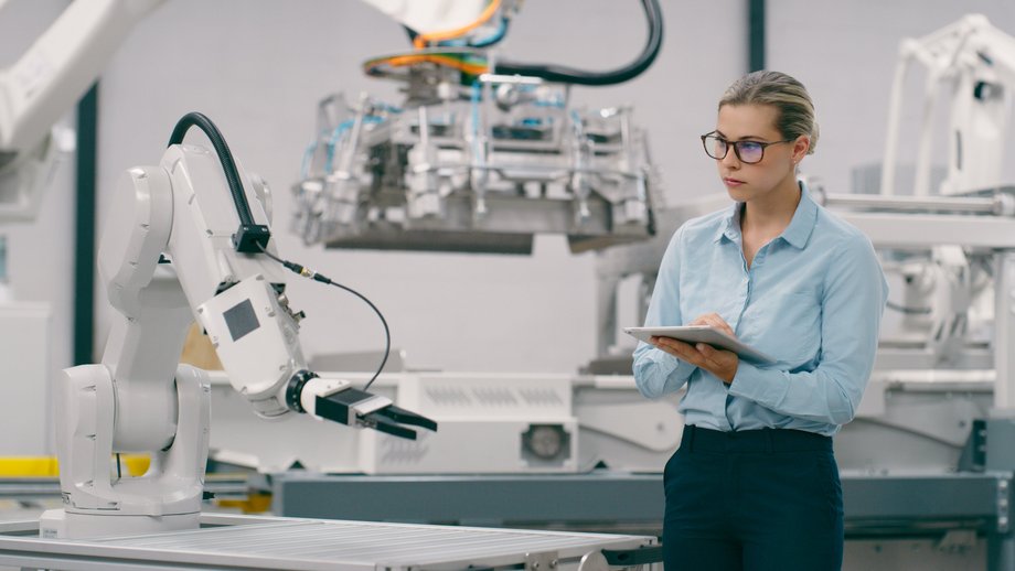 A female engineer monitors a robotic arm within a modern factory setting while holding a digital tablet.