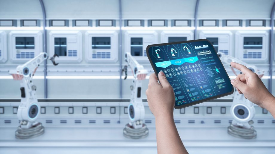 Worker monitors yellow robot arms with a tablet, sparks flying in an industrial hall