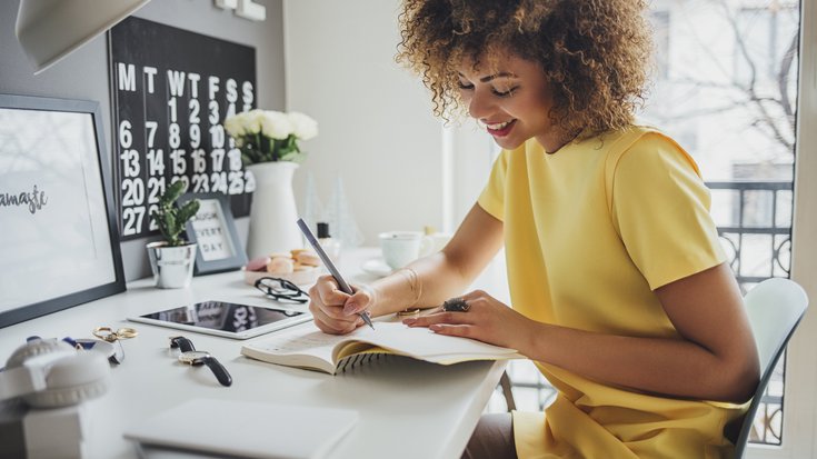 A laughing woman sits at a desk and writes something down in a notebook