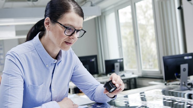 A woman sits at a workstation with a magnifying glass in her hand