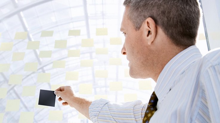 A businessman holds up a black post-it in front of a wall with many yellow post-its